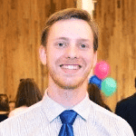 Professional headshot of man with reddish hair wearing blue tie and light colored shirt smiling at camera