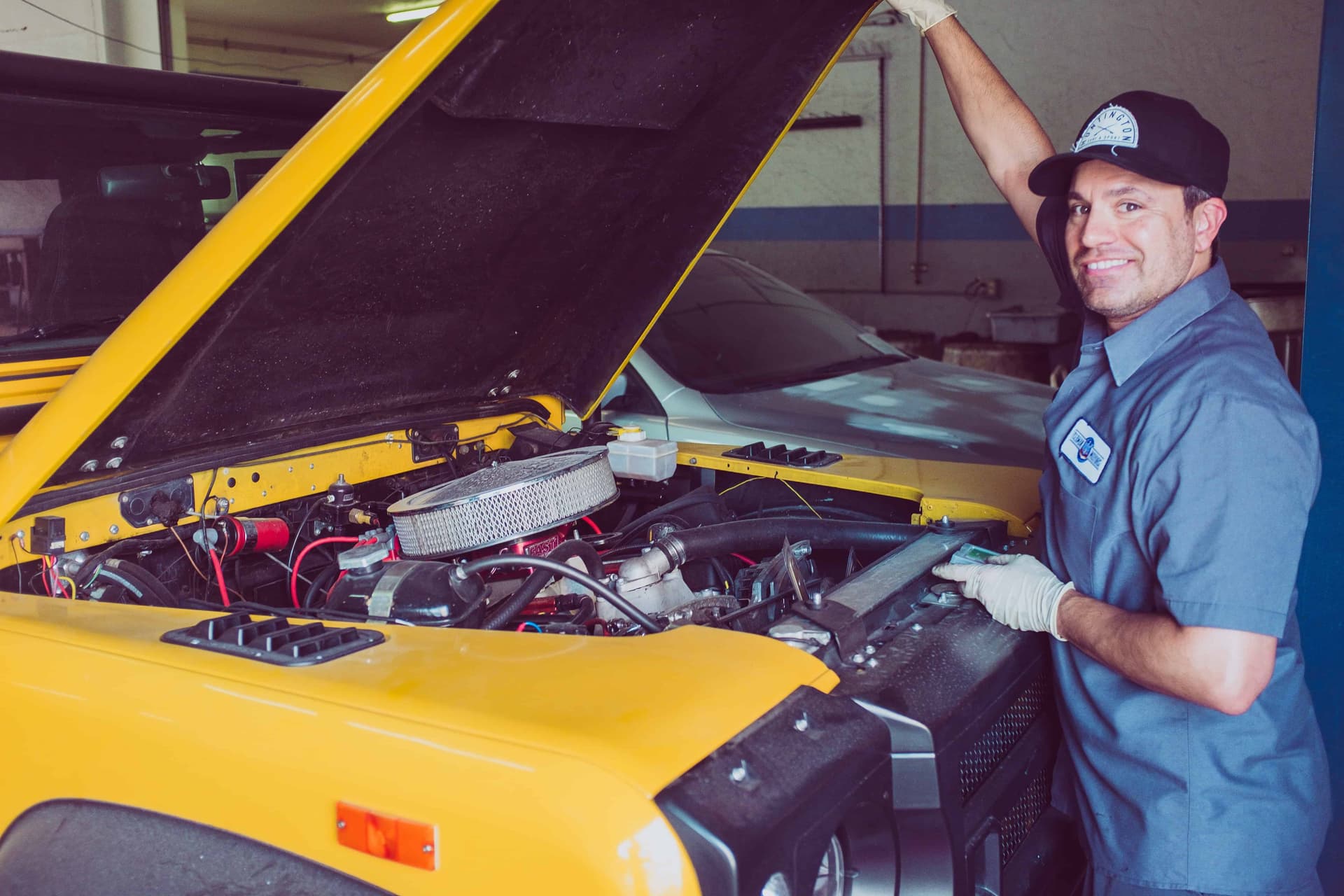 Friendly mechanic in uniform and cap with open hood on yellow vehicle in shop