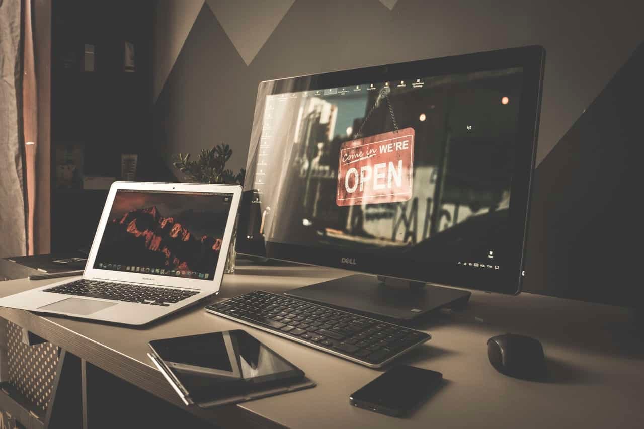 Desk with laptop, tablet, and phone; background monitor shows a Come In We're Open sign