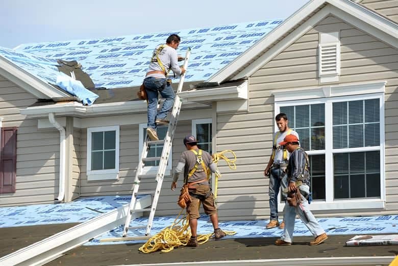 Four roofers install blue synthetic underlayment on a house roof, one working from a ladder