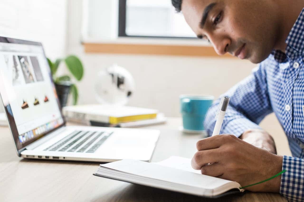 Man in a checkered shirt writing in a notebook beside a laptop showing a website