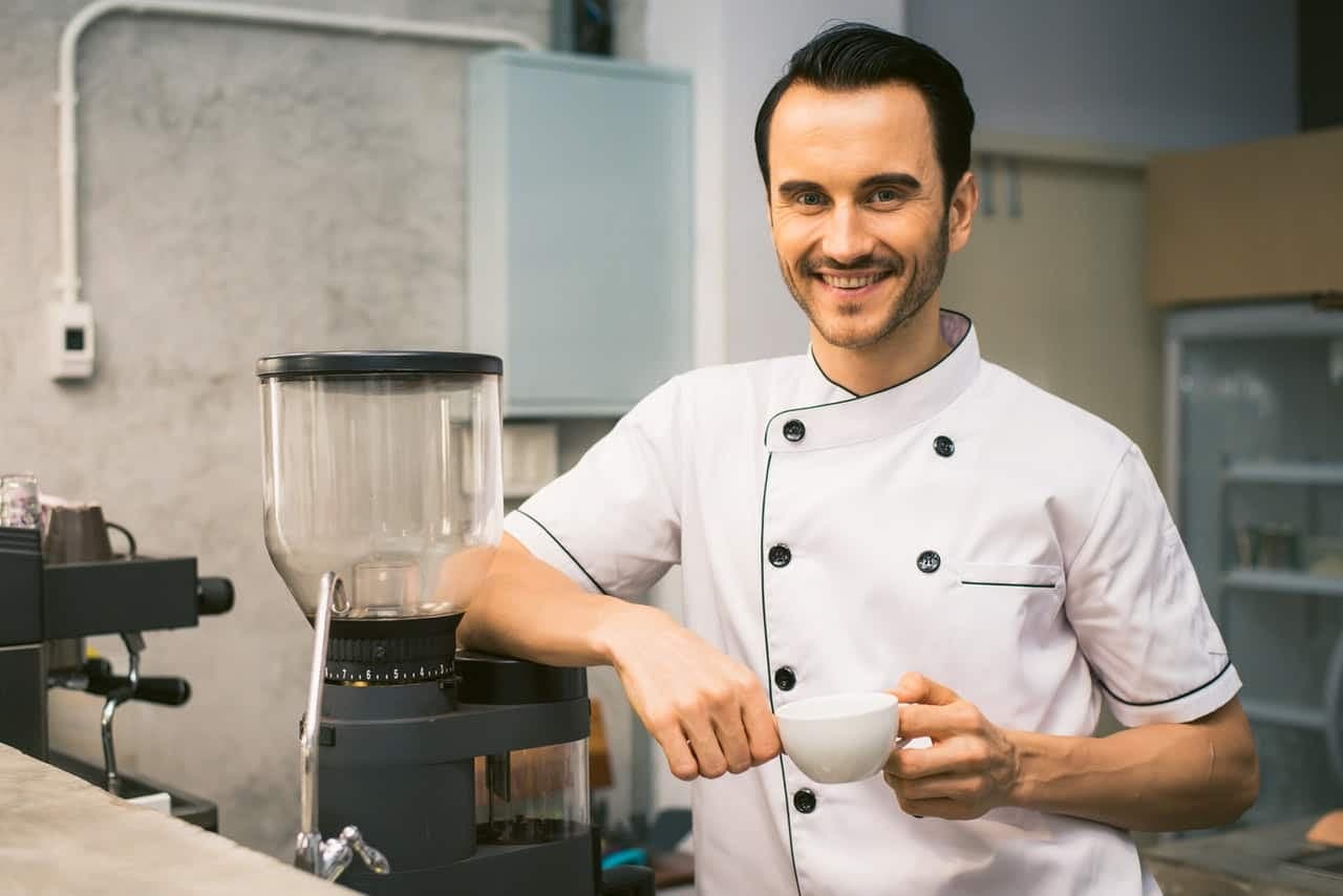 Chef in white coat smiling with coffee near professional kitchen equipment