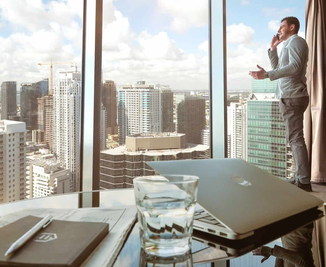 Man in a blazer smiling on a phone call, gesturing near floor-to-ceiling windows above a city skyline