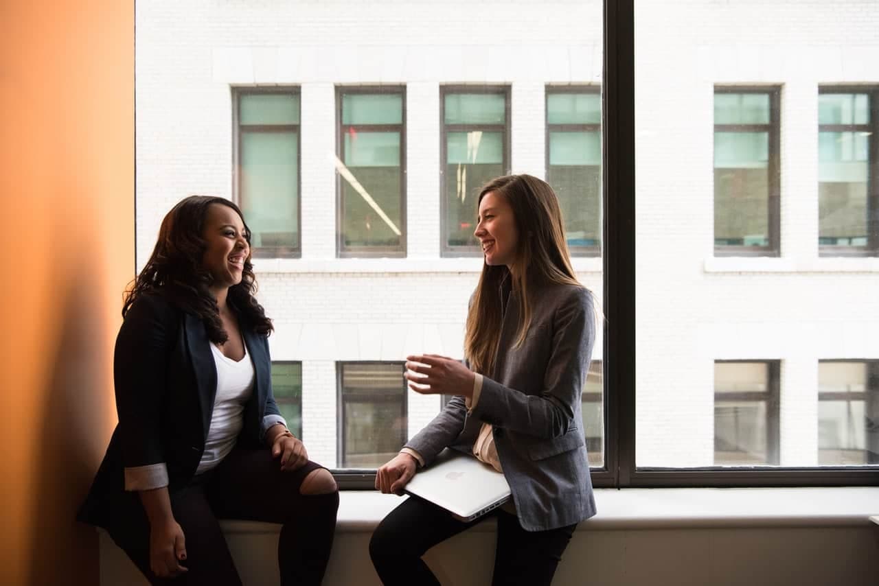 Two women talking by a large window, one in a dark blazer, the other in a gray blazer holding a laptop on her lap