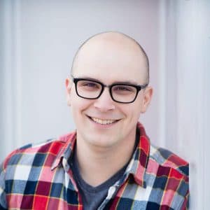 Professional headshot of bald man wearing black-framed glasses and red and blue plaid shirt, smiling