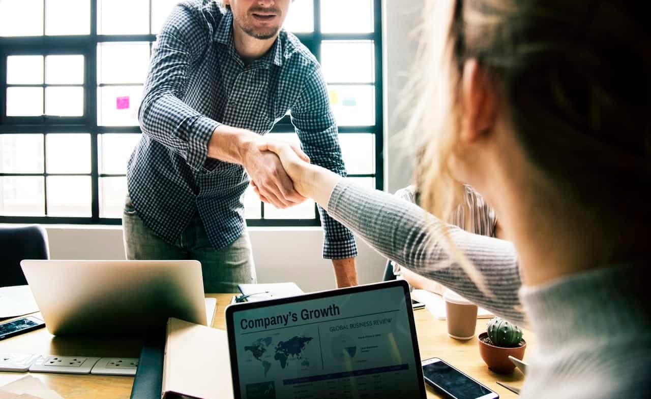Two professionals shaking hands at desk with business growth materials and laptop