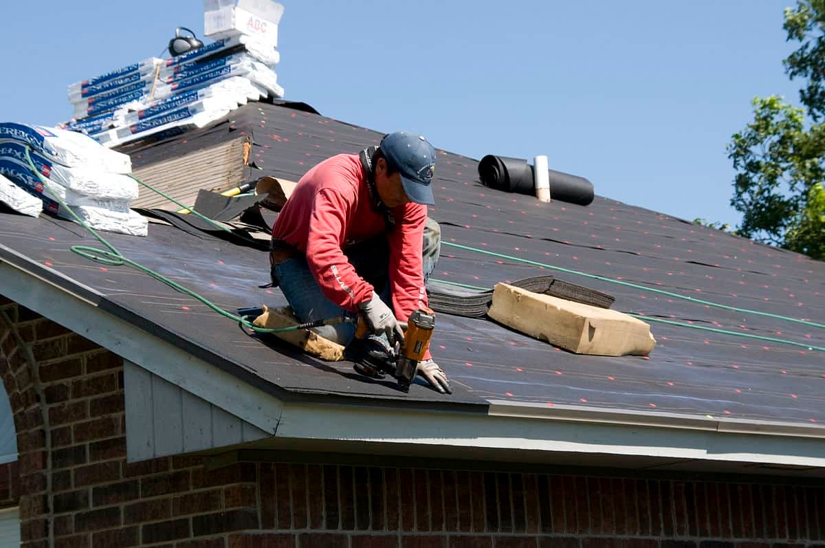 Roofer in a red shirt nail-gunning shingles on a roof covered in black felt underlayment