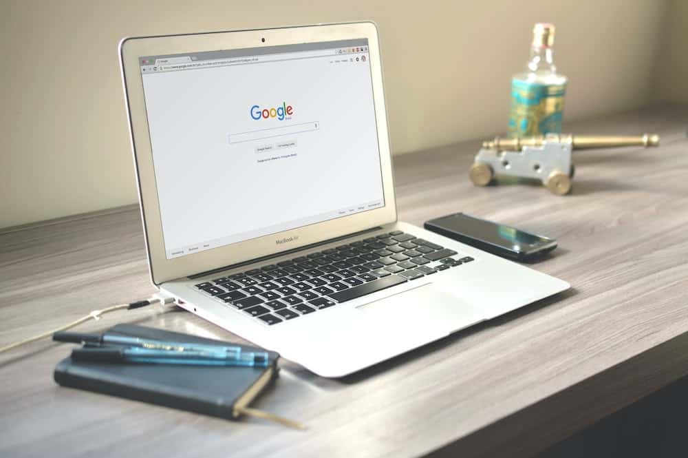 MacBook with Google homepage on wooden desk with smartphone and accessories