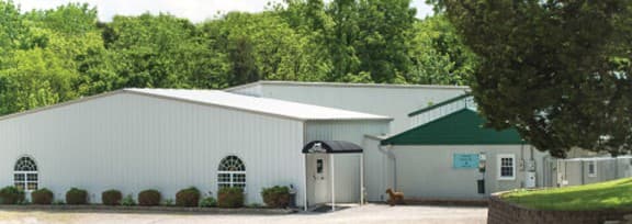 White metal building with a dark green triangular gable end, one arched window, and a black door awning, surrounded by trees