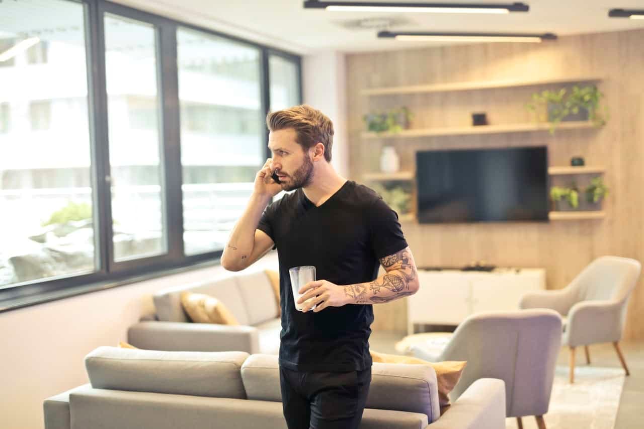 Man on phone call in modern living room with couch and shelving while holding a drink