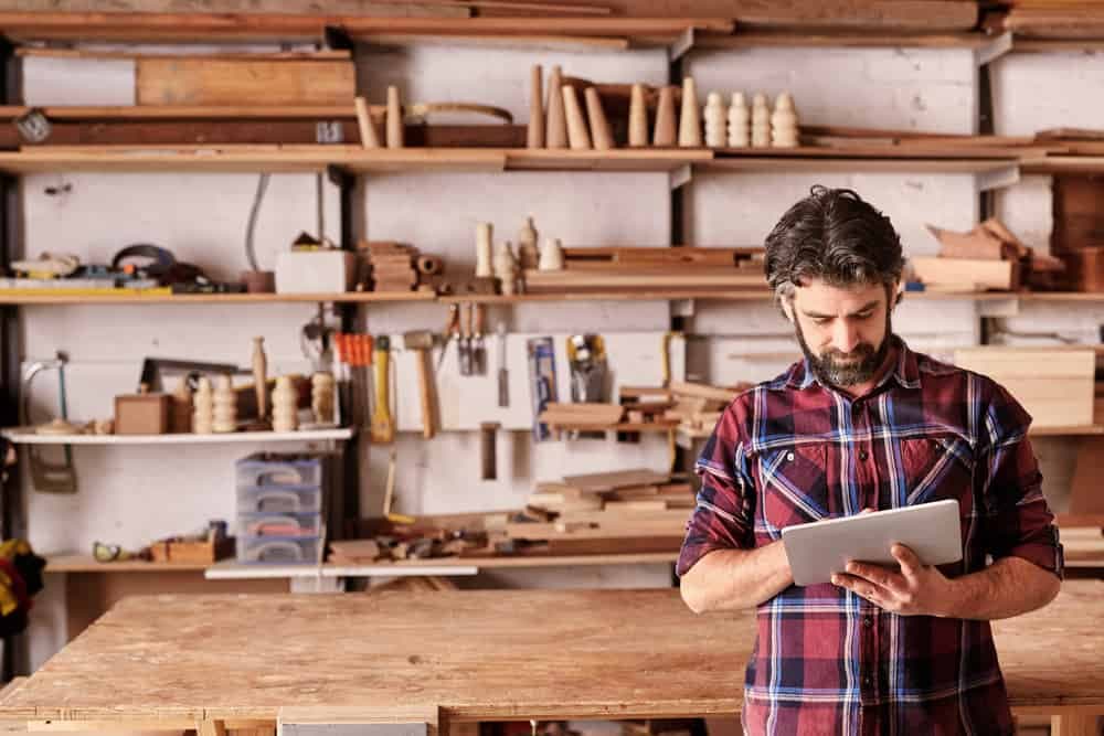 Workshop owner in plaid shirt reviewing tablet with tools and shelves behind
