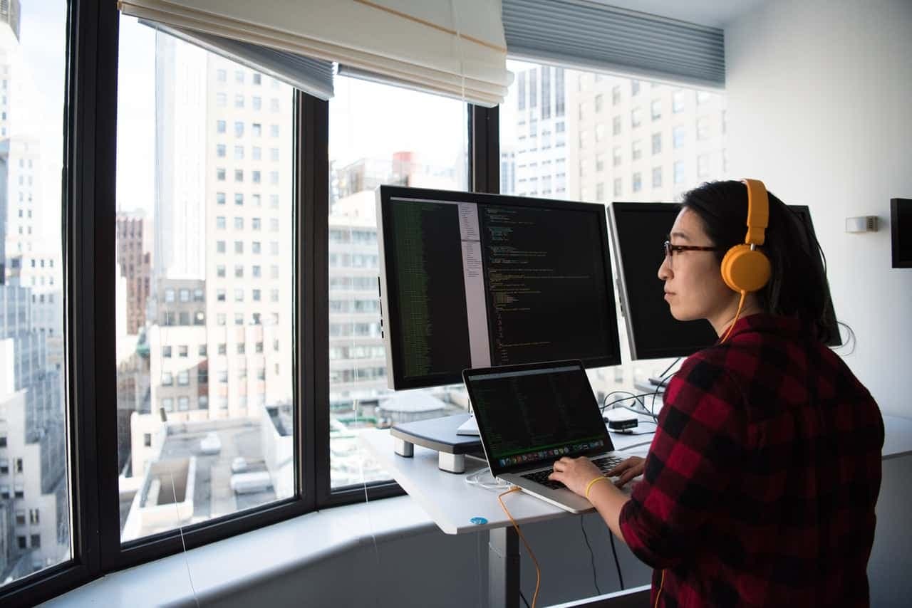 Developer at desk with yellow headphones working on laptop and dual monitors overlooking city