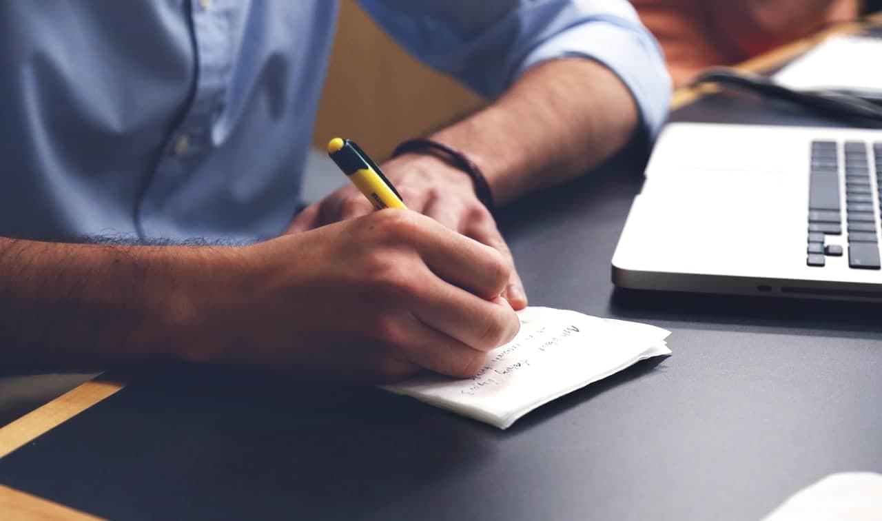 Close-up of hands writing on notepad with pen, person in blue shirt with laptop visible in background