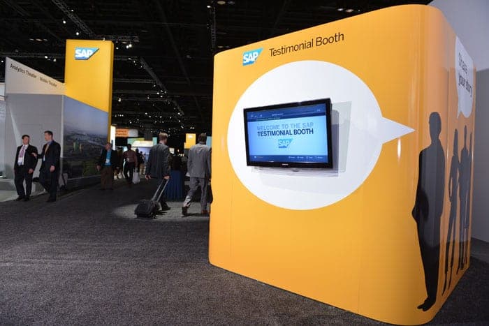 Yellow SAP testimonial booth at conference with screen display and silhouettes, conference attendees in background