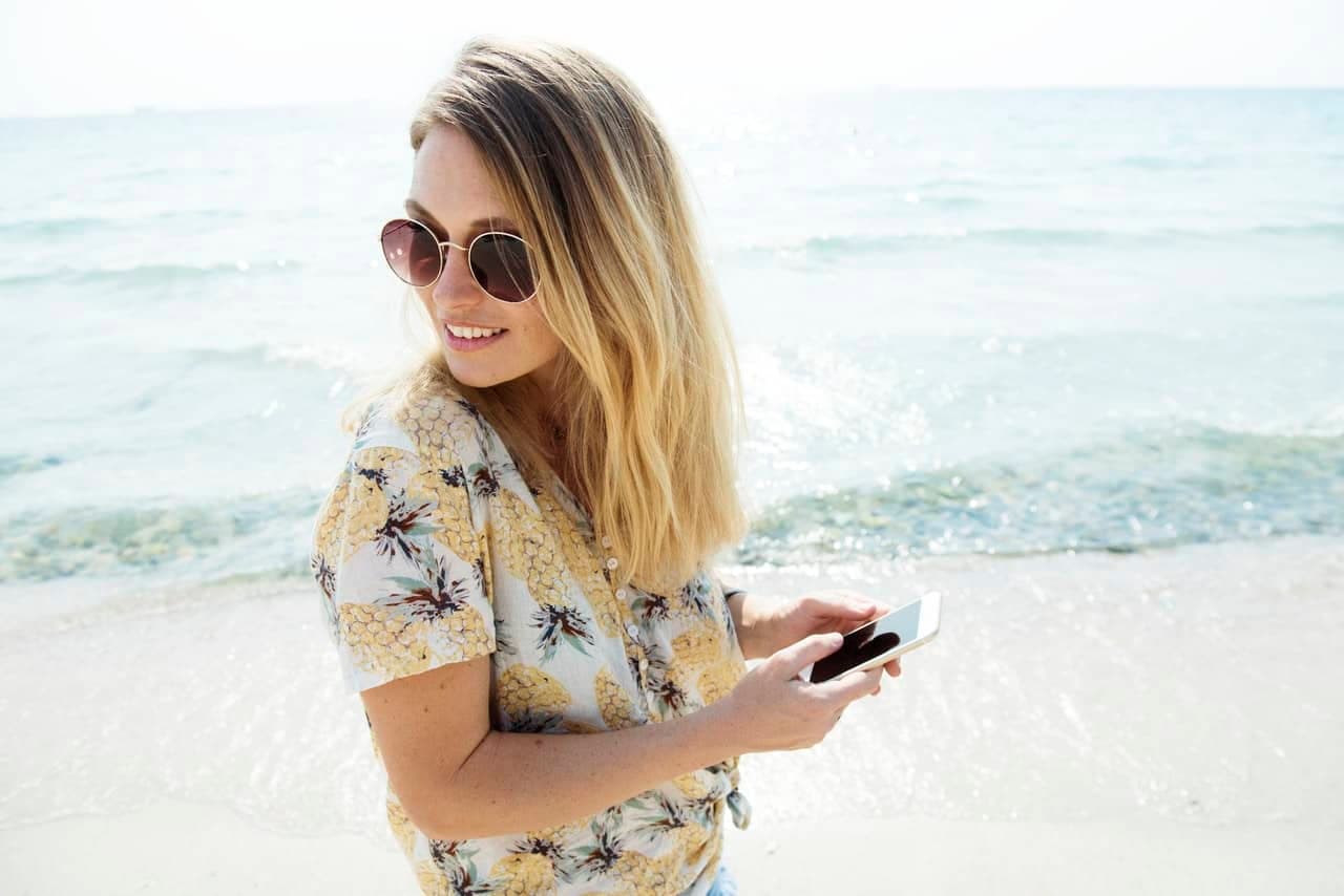 Woman on beach wearing sunglasses and floral shirt holding phone looking at ocean