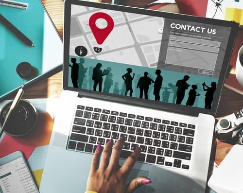 Laptop displaying contact form with map and people silhouettes, hand on keyboard with colorful desk accessories