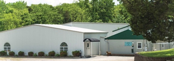 White metal building with a dark green triangular gable end, one arched window, and a black door awning, surrounded by trees