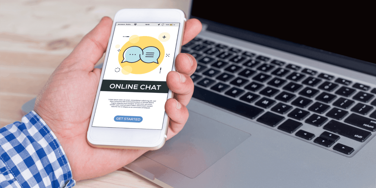 Hand holding phone displaying online chat interface with message bubbles icon, near laptop