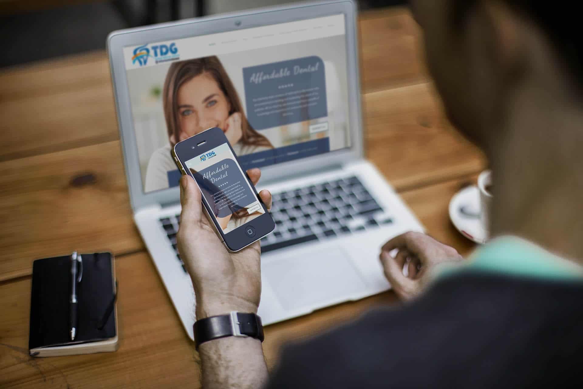 Hand holding phone displaying dental practice website with laptop background