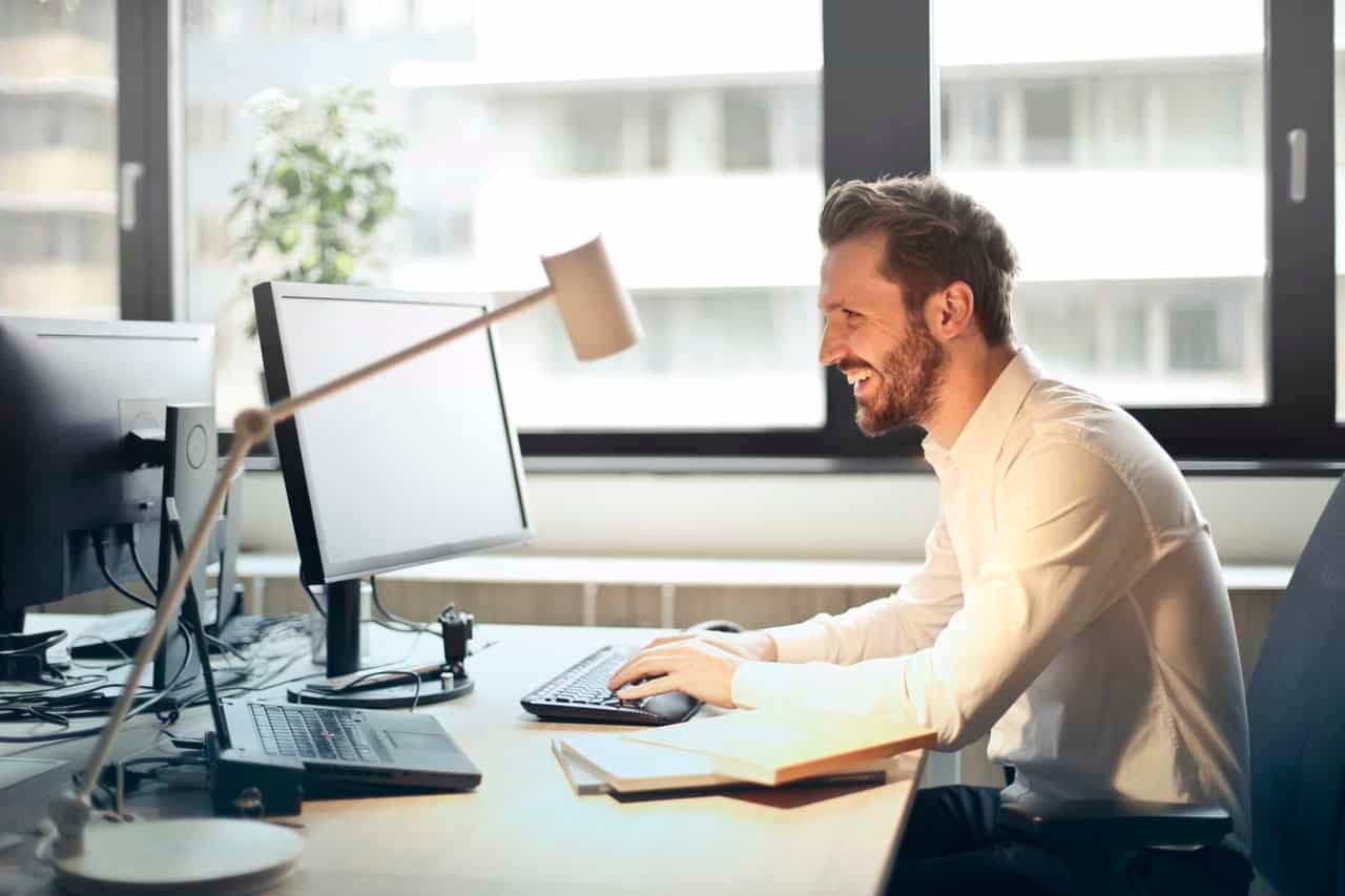 Man working at desk with dual monitors in bright high-rise office overlooking city skyline