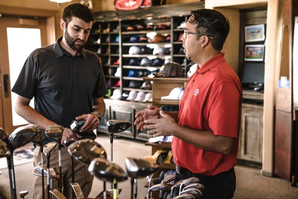 Retail salesman in red shirt discussing golf equipment with customer in pro shop