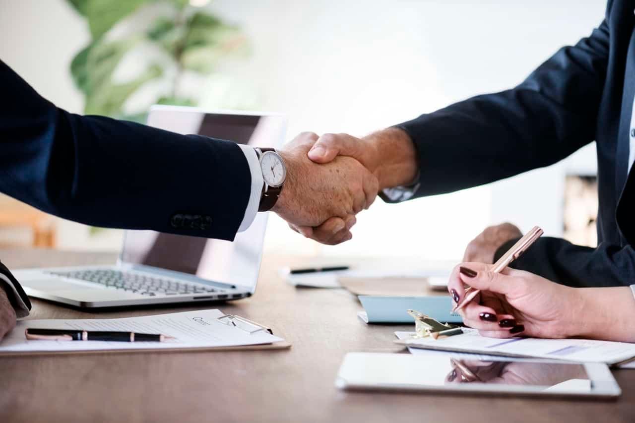 Two business professionals handshake over desk with documents and laptop