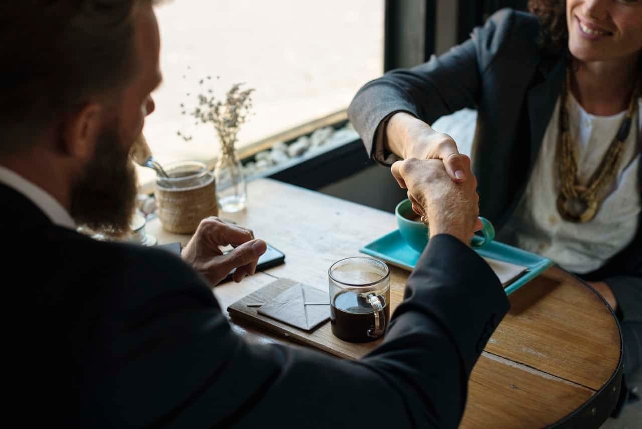Man and woman shaking hands across a café table set with coffee and a small flower vase