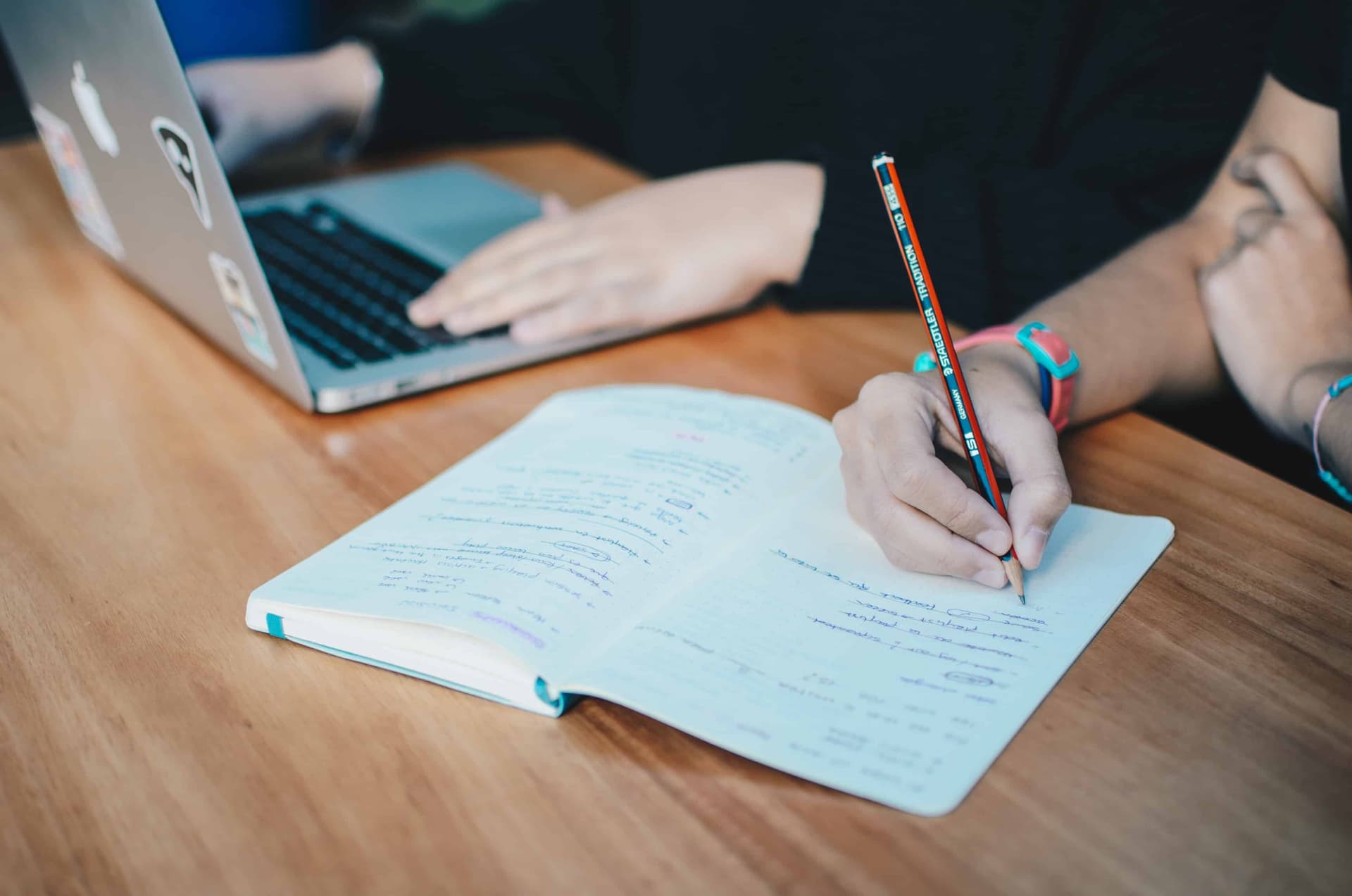 Person writing in light blue notebook at wooden desk with laptop in background