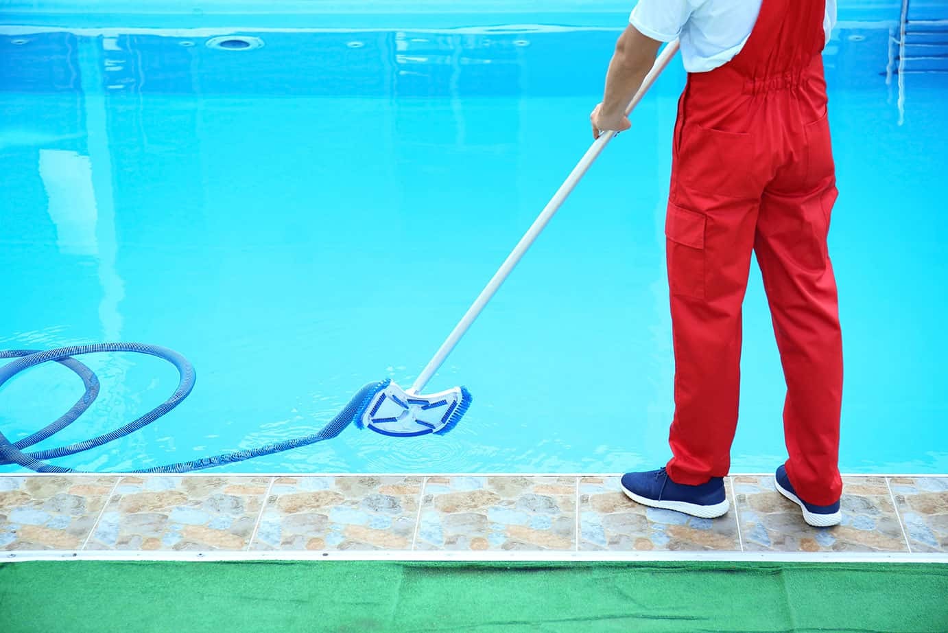 Pool worker in red overalls vacuuming the pool floor with a hose-connected brush head