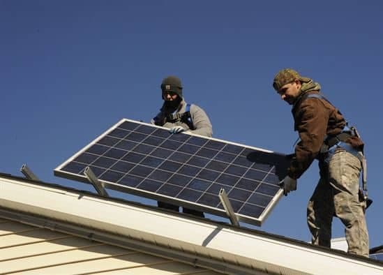Two workers installing a solar panel on residential rooftop against clear blue sky