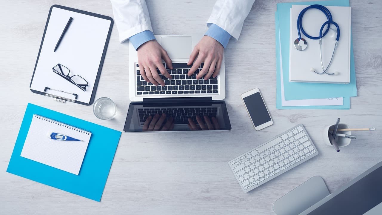 Doctor's hands typing on a laptop, with a stethoscope, keyboard, and desk supplies nearby